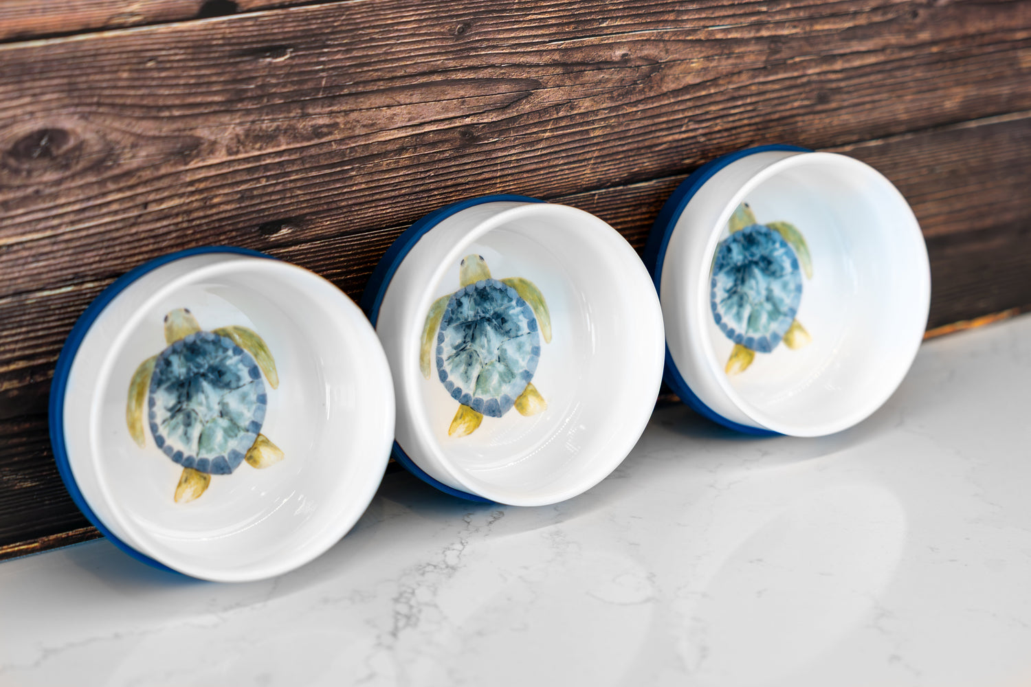 Three white ceramic kids bowls with blue sea turtle illustrations and blue silicone grips, lined up on a marble surface with wood background — safe, non-toxic dinnerware alternative to plastic plates for kids.