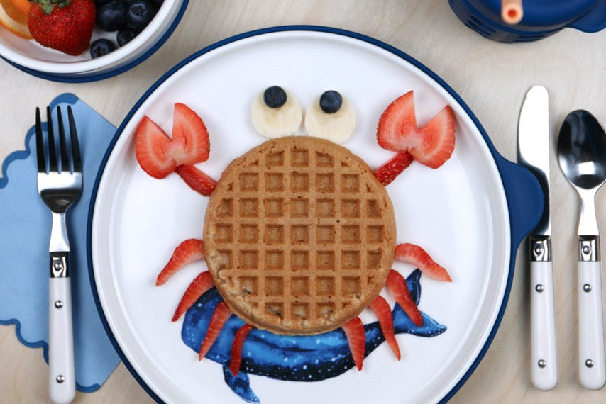 Crab-shaped waffle on a whale shark plate with fruit and cutlery on a wooden table