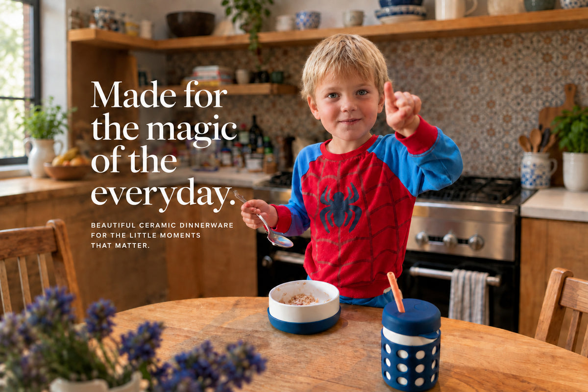 A young child wearing a red and blue pajama shirt stands at a wooden table in a kitchen, holding a spoon with ceramic kid's plates on the table.